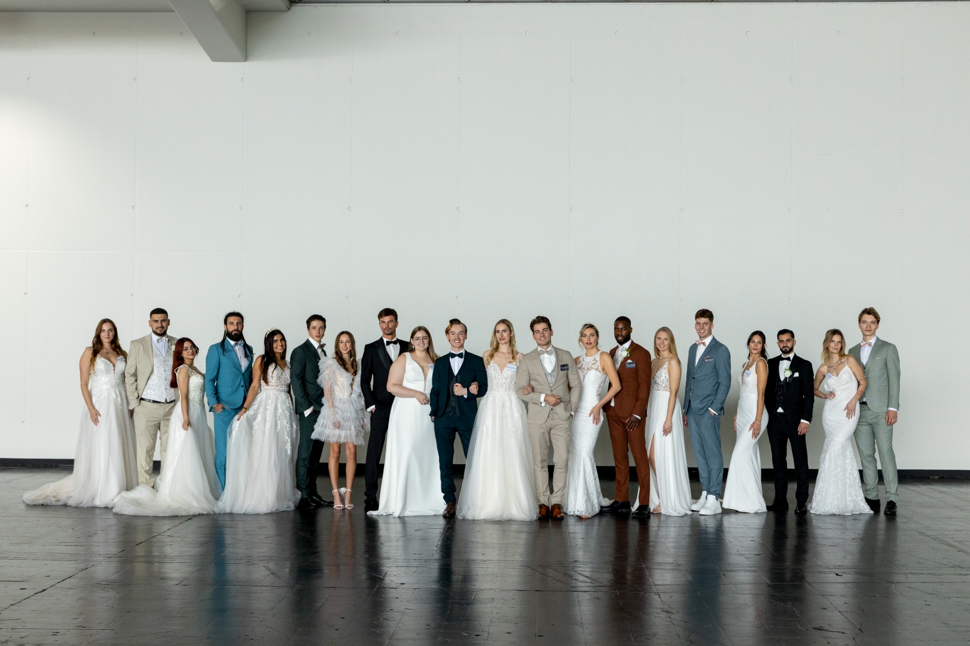 Eine Gruppe von Hochzeitsgästen in Braut- und Anzugkleidung vor einer weißen Wand.