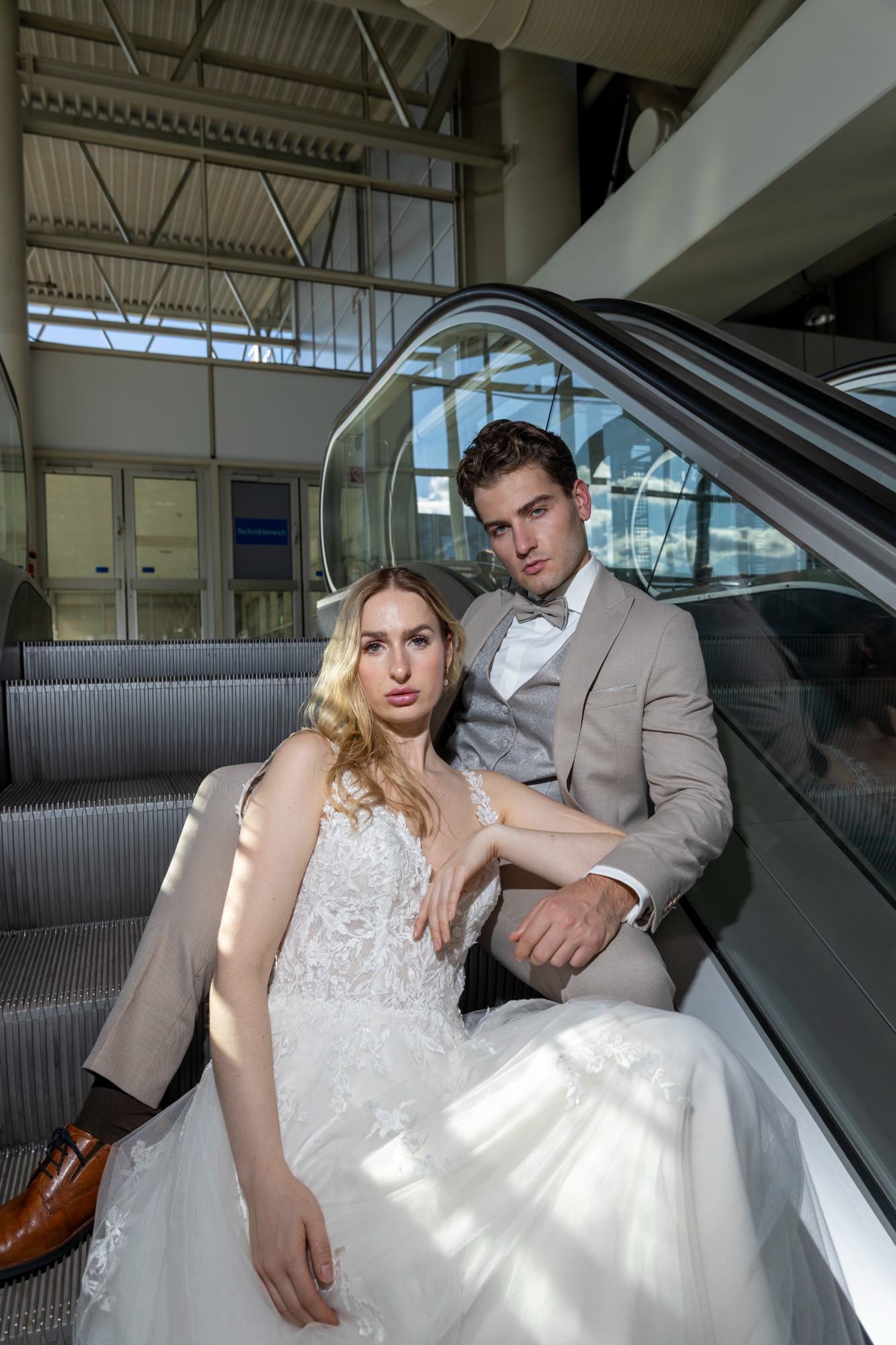 Braut und Bräutigam posieren elegant auf einer Rolltreppe in hellem, modernem Gebäude.