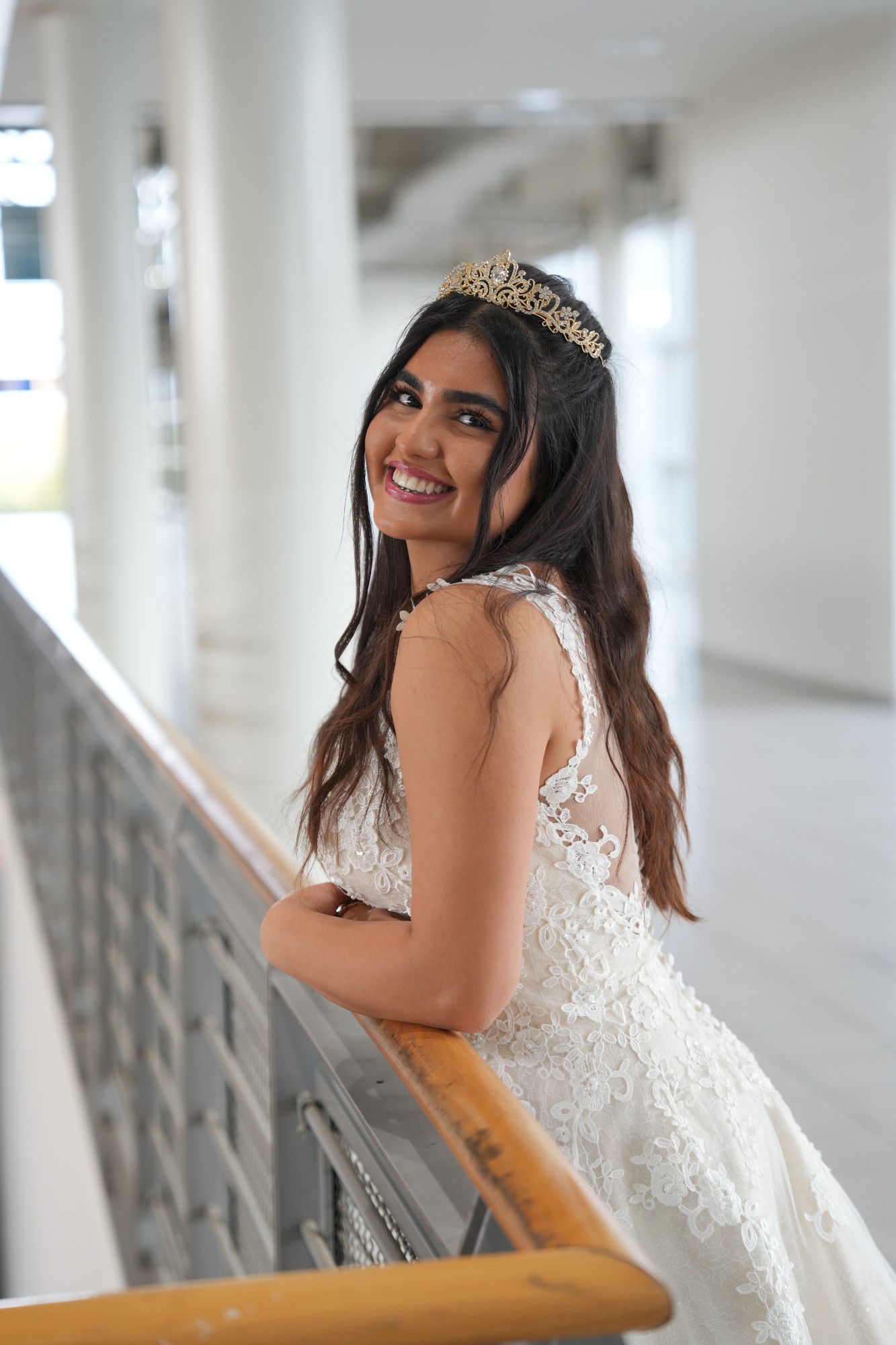 Hochzeitsbraut lächelt elegant in einem festlichen Kleid, vor einer hellen, modernen Kulisse.