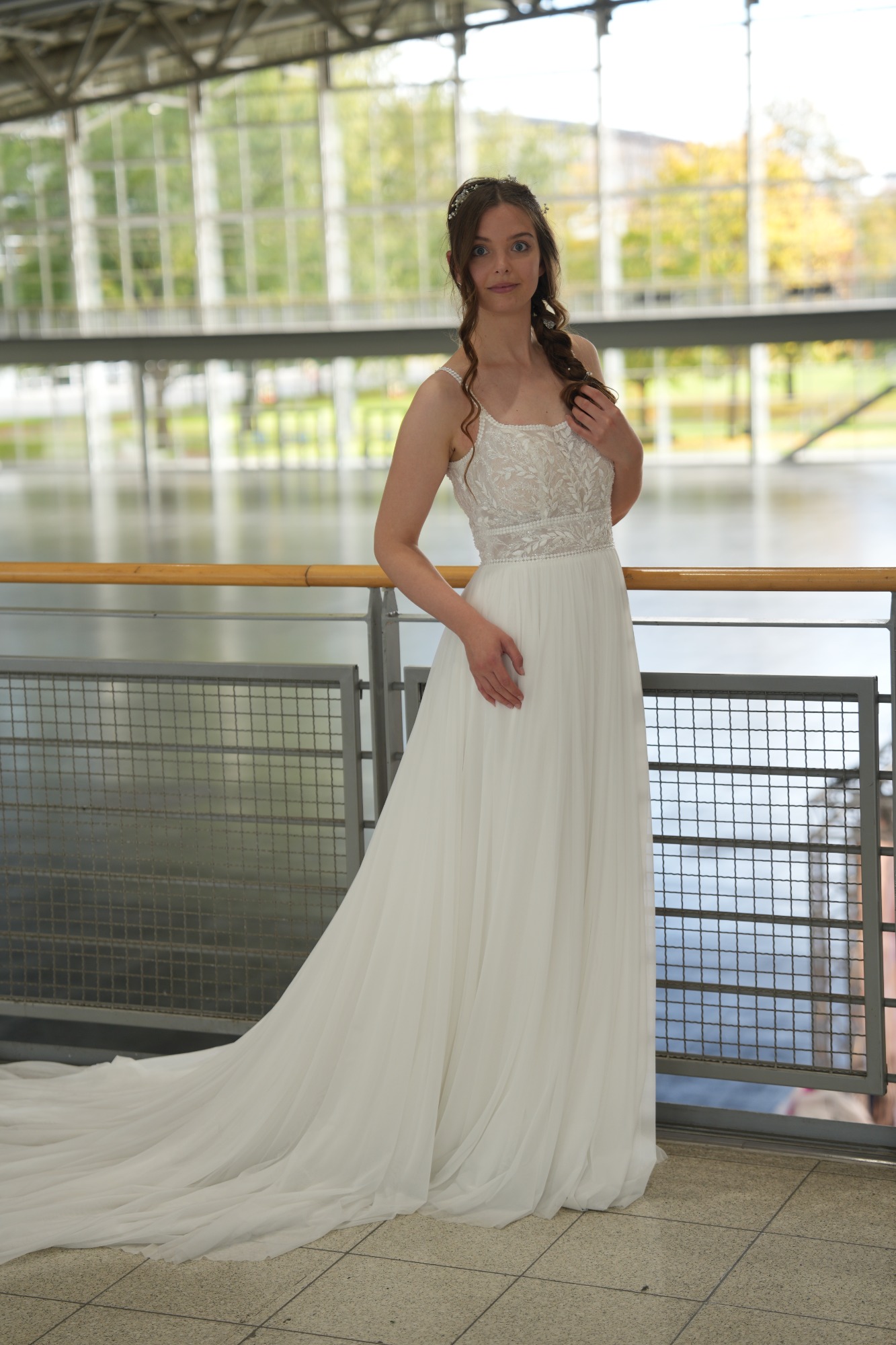 Braut in einem weißen Hochzeitskleid steht vor einem großen Fenster.