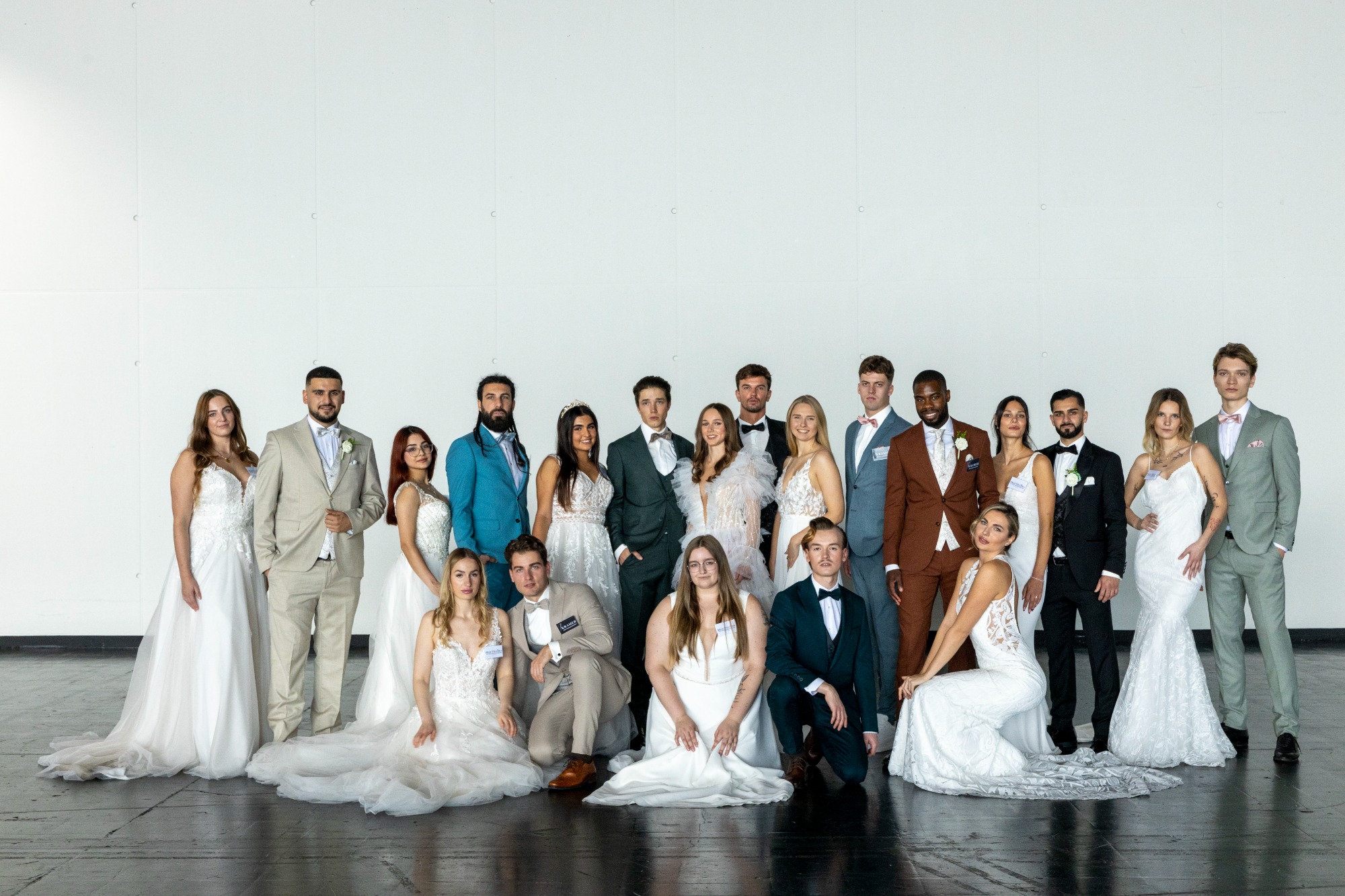 Gruppenfoto von 20 Personen in festlicher Kleidung, überwiegend in Weiß, vor einfarbigem Hintergrund.