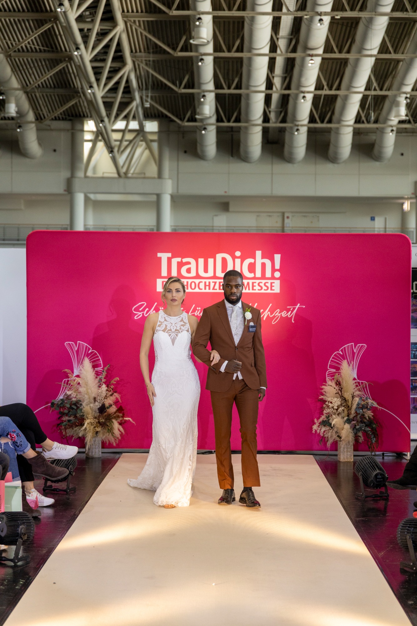 Braut und Bräutigam in eleganter Hochzeitsmode auf einem Laufsteg mit pinkem Hintergrund.