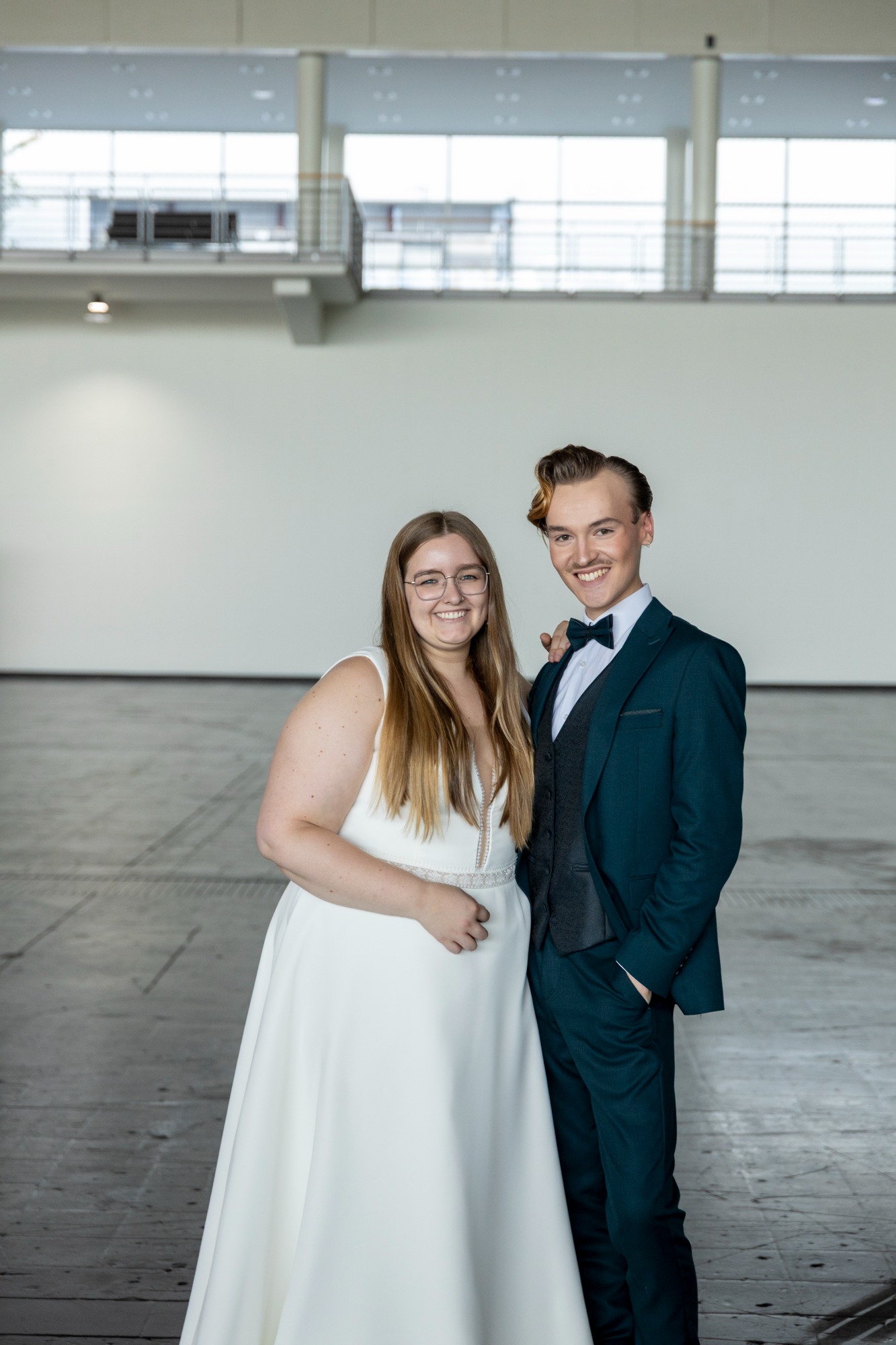 Brautpaar in eleganter Hochzeitskleidung lächelt in einem modernen, hellen Raum.