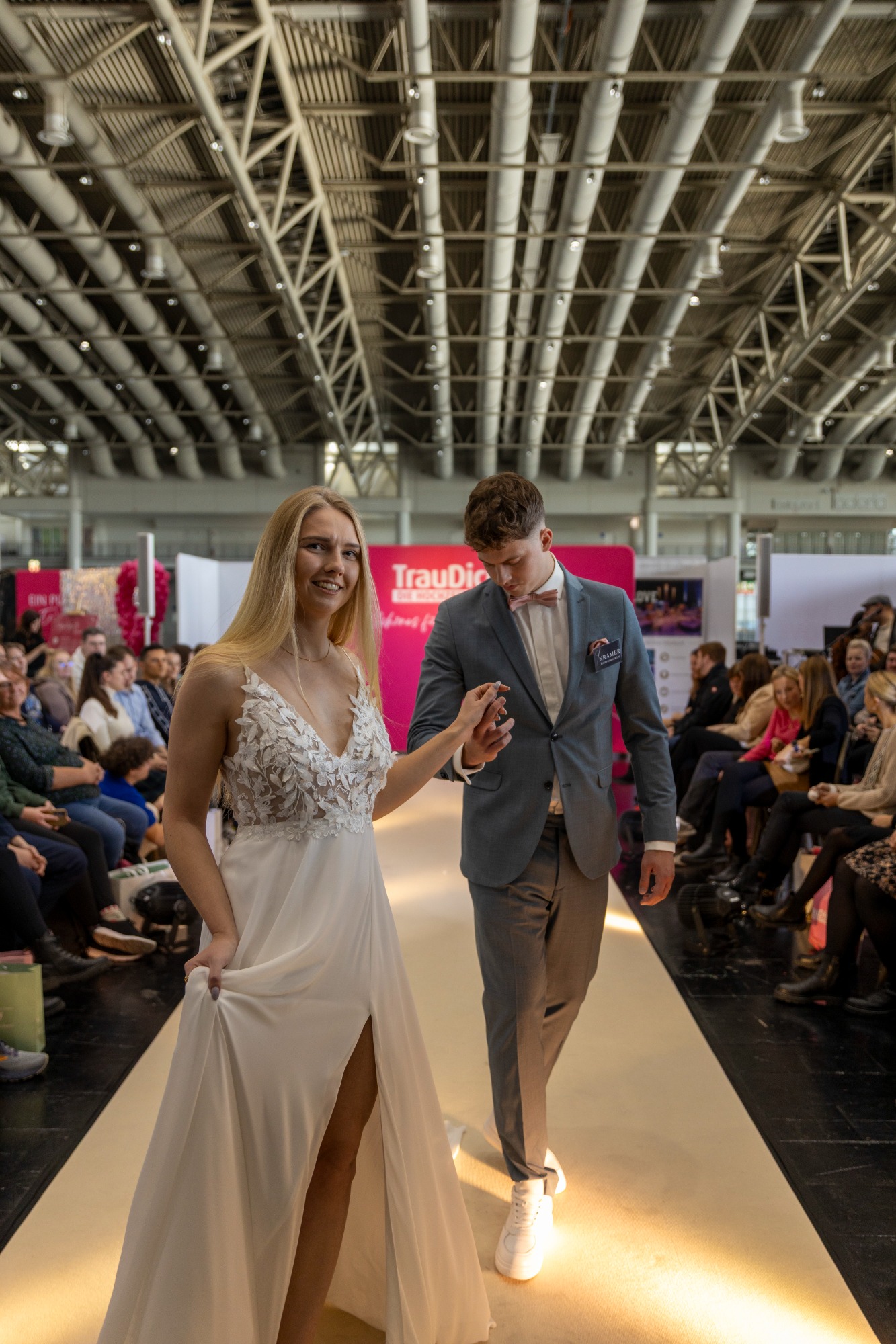 Model in weißem Kleid und Model in Anzug gehen über den Laufsteg bei einer Modenschau.