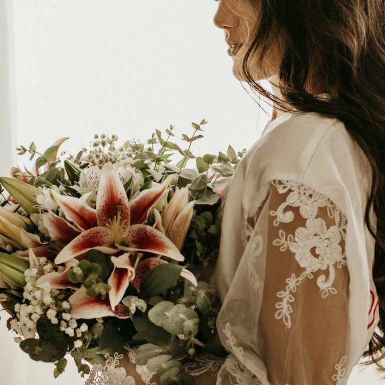 Braut-BrautBlumen-klassisch-Brautkleid-Haury-weiß Frau mit langen Haaren hält einen üppigen Blumenstrauß, darunter Lilien und grüne Zweige.