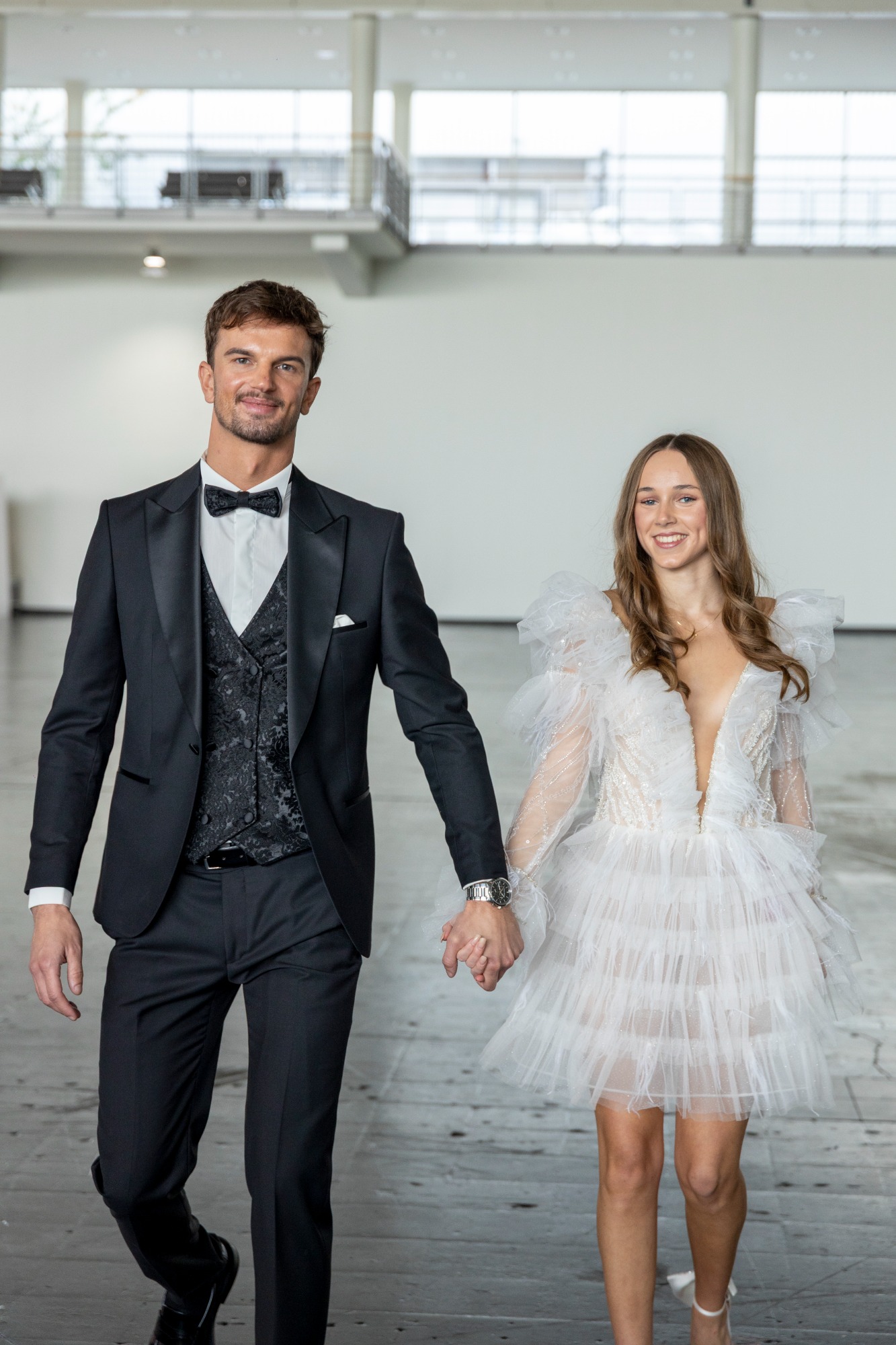 Brautpaar in eleganter Kleidung, lächelnd, Hand in Hand in einem modernen Raum.