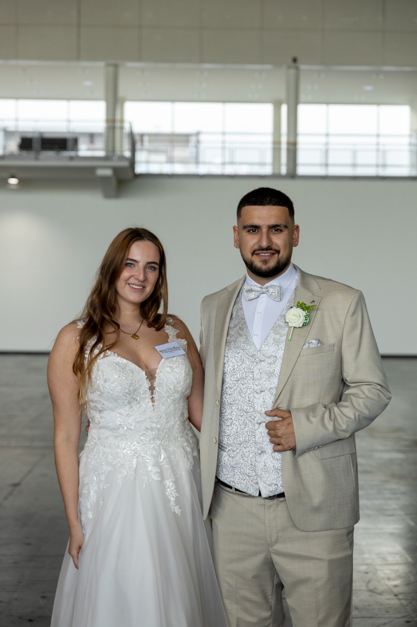 Braut und Bräutigam in eleganter Hochzeitskleidung, lächelnd in einem großen Raum.