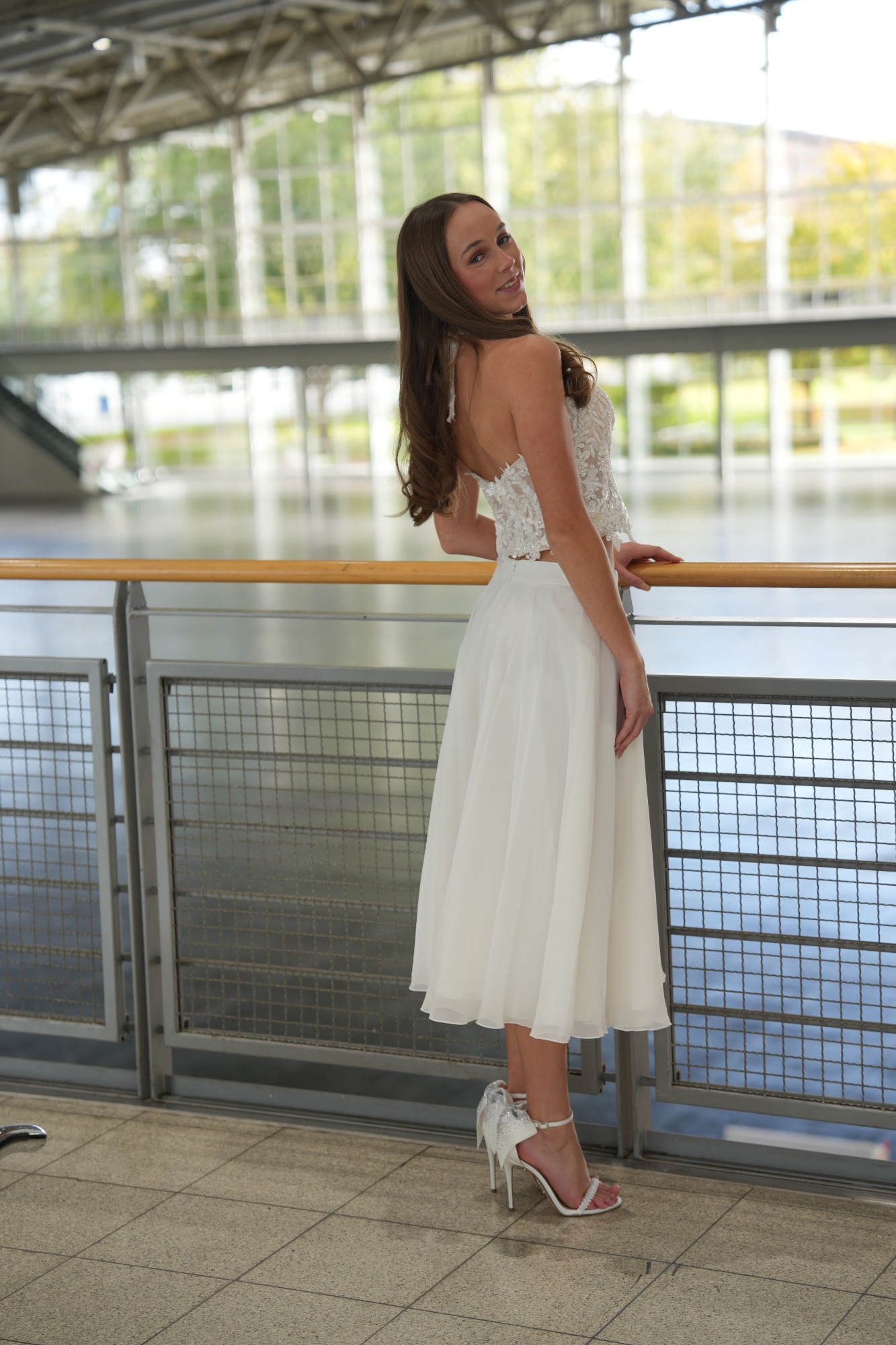 Frau in weißem Kleid steht an einem Geländer mit Glasarchitektur im Hintergrund.