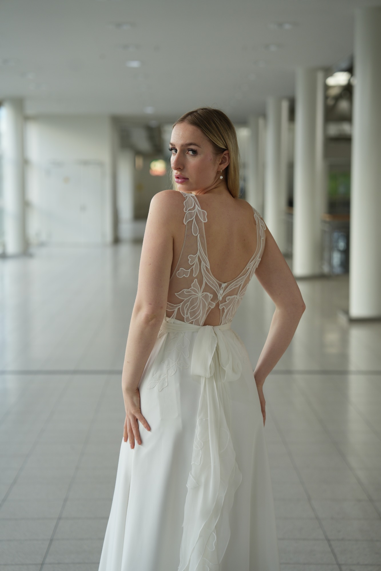 Frau in einem weißen Kleid mit zarter Rückendetails, steht in einem hellen Raum.