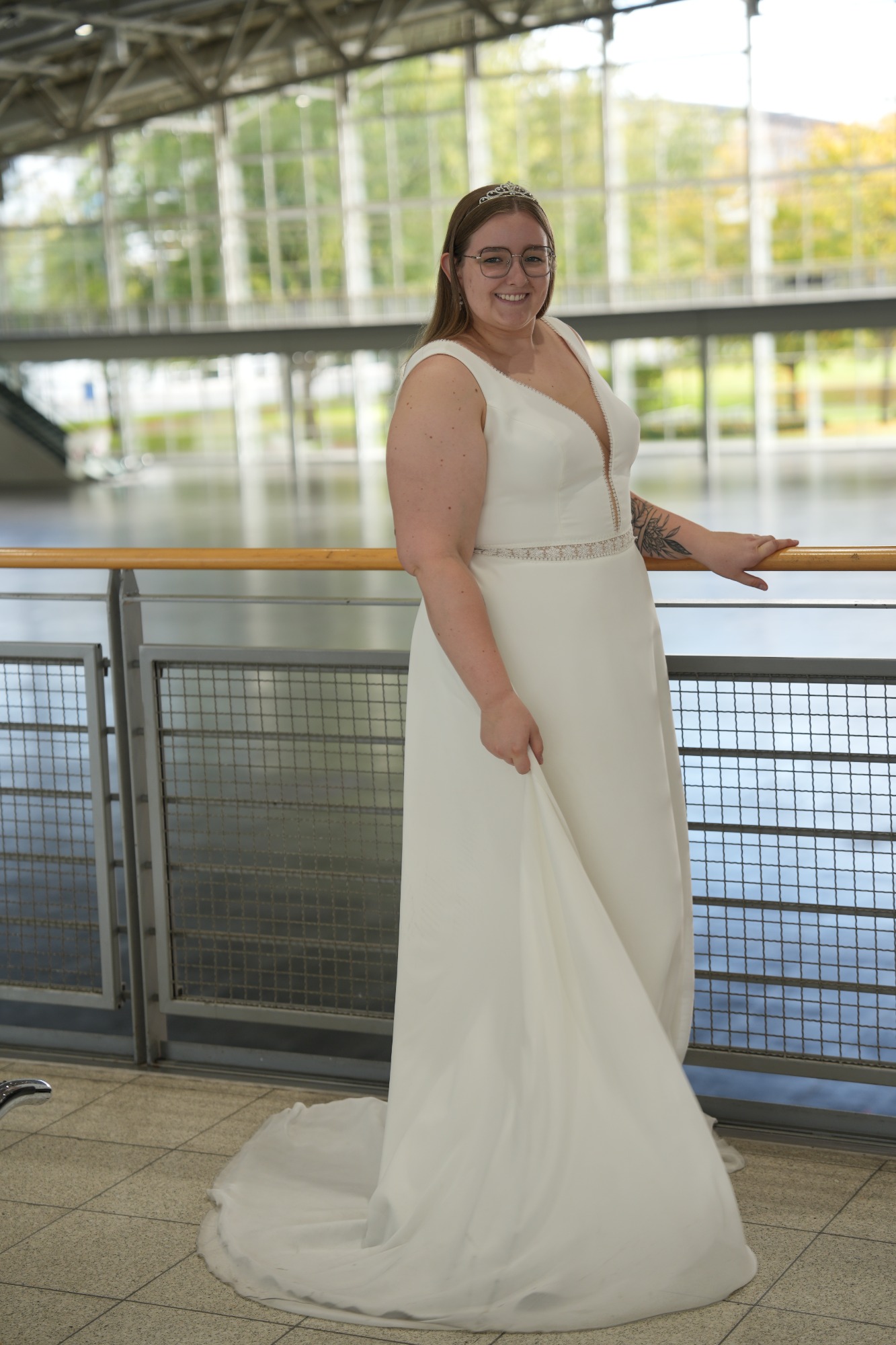 Eine Frau in einem eleganten, trägerlosen Hochzeitskleid steht am Fenster.
